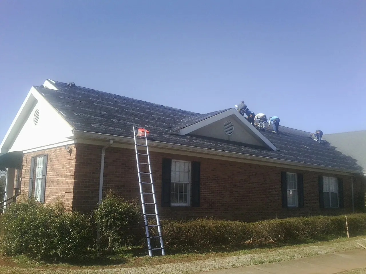 Crew tearing shingles off a brick ranch roof with a ladder set — Emergency Roof Repair work by our crew in East Ryegate