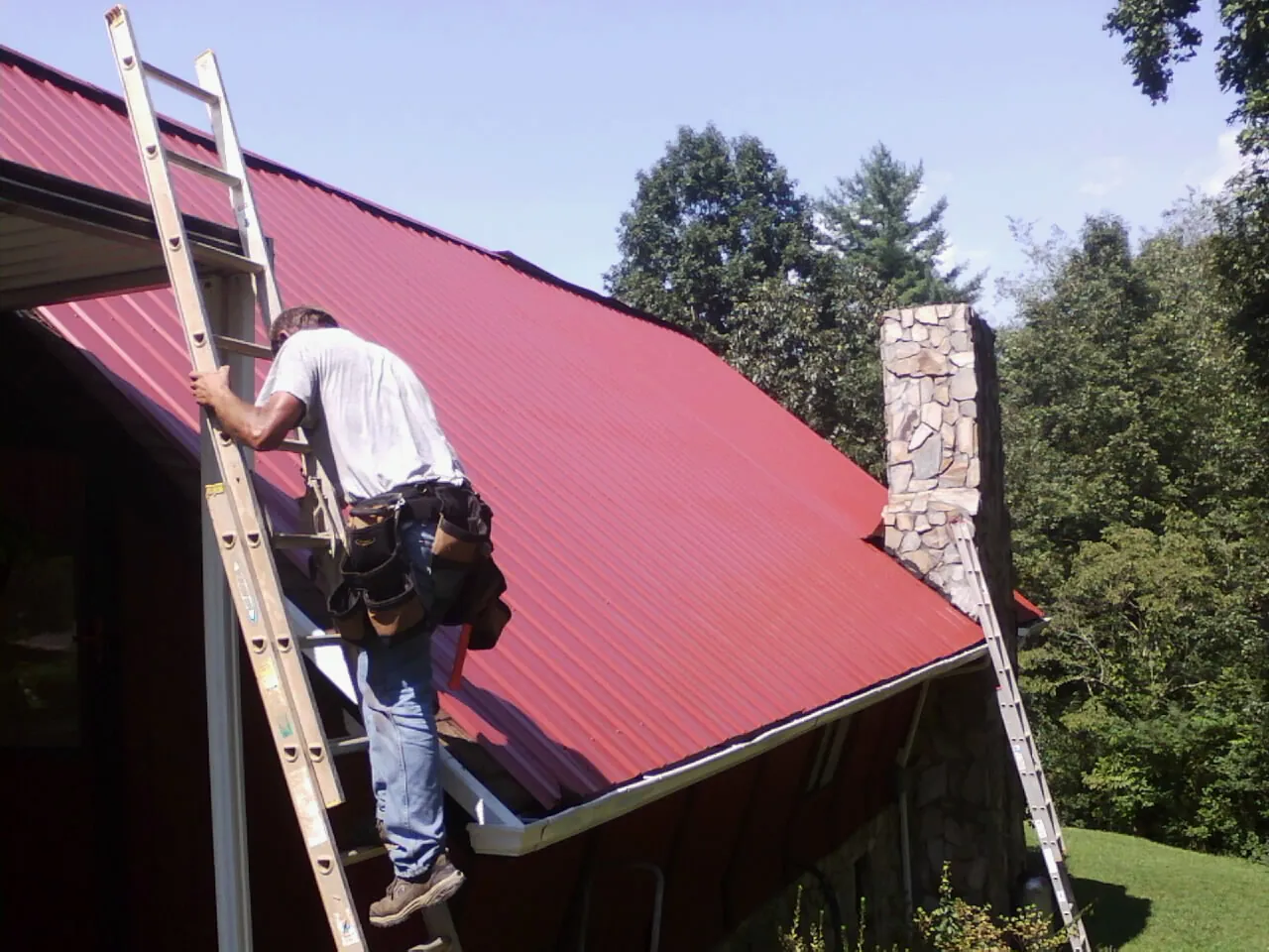 Roofer on a ladder beside a red metal roof and stone chimney — Soffit & Fascia Repair by Roofing Brothers in East Ryegate