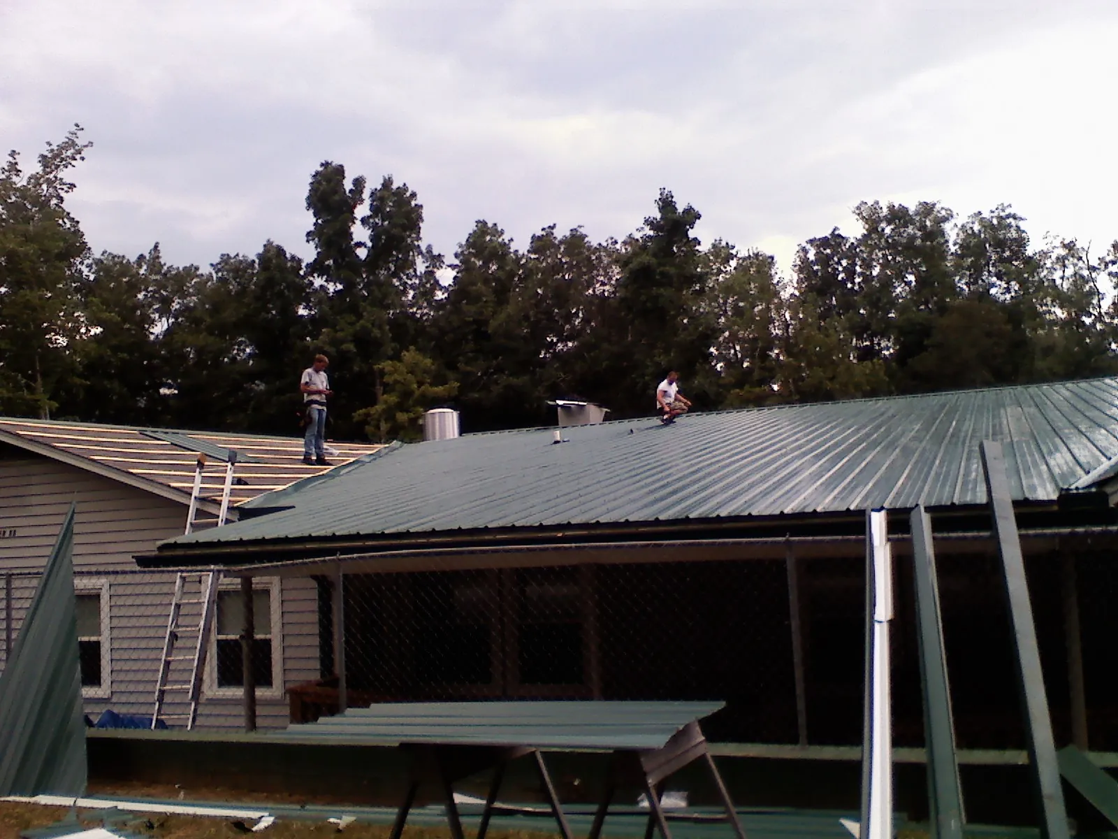 Two roofers installing green metal panels above a covered porch in East Ryegate